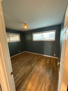 Doorway view into a bedroom painted dark teal with white window trim, brass light fixture and oak flooring