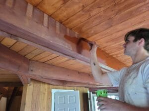 Painter brushing stain onto an exposed wood ceiling beam inside a cabin-style room