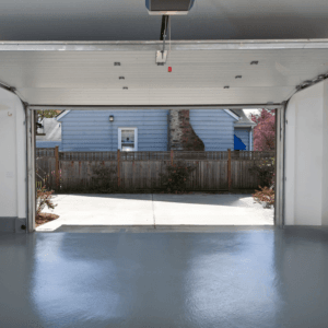 Empty garage with a solid gray painted concrete floor and open door facing a blue house