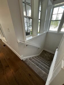 Stairwell freshly painted white with white handrail, window trim and baseboards leading to carpeted stairs