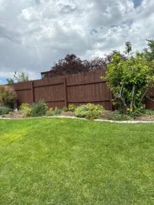 Backyard wood fence coated in solid dark brown stain behind a flower bed and green lawn