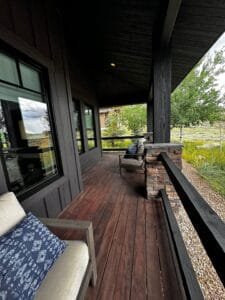 Covered porch with dark reddish hardwood decking, black stained siding, beams and railing