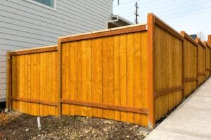 Newly built cedar fence with golden stain and darker trimmed posts beside a gray house