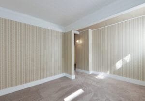 Empty room with dated cream floral striped wallpaper, white beadboard ceiling and beige carpet