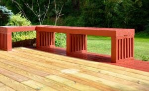 Deck staining in progress with bare pine boards in front of a red stained section and benches