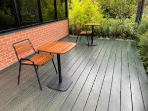 Dark green-gray deck with teak bistro tables and chairs beside a red brick wall
