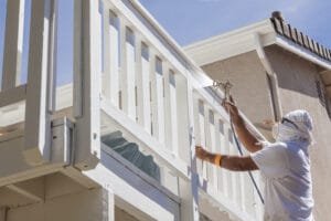 Painter in protective gear spray painting a white wooden balcony railing on a stucco home