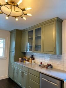 Kitchen cabinets painted olive green with glass-front uppers, white stacked tile backsplash and gold light fixture