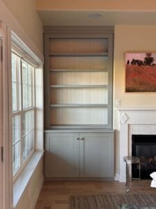 Built-in bookcase painted soft gray-green with white shiplap back beside a white fireplace mantel
