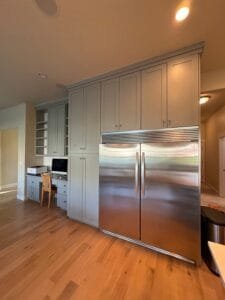 Tall pantry and over-fridge cabinets painted gray-green around a stainless built-in fridge beside a desk nook